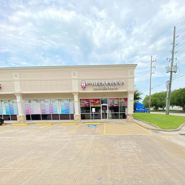 A storefront of "La Michoacana Autentica" with a clear sky, visible parking spaces, and large windows displaying colorful posters.