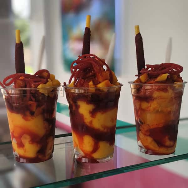 Three cups filled with chopped mango, drizzled with a syrup, topped with a tamarind stick, and garnished with spicy candy strips.