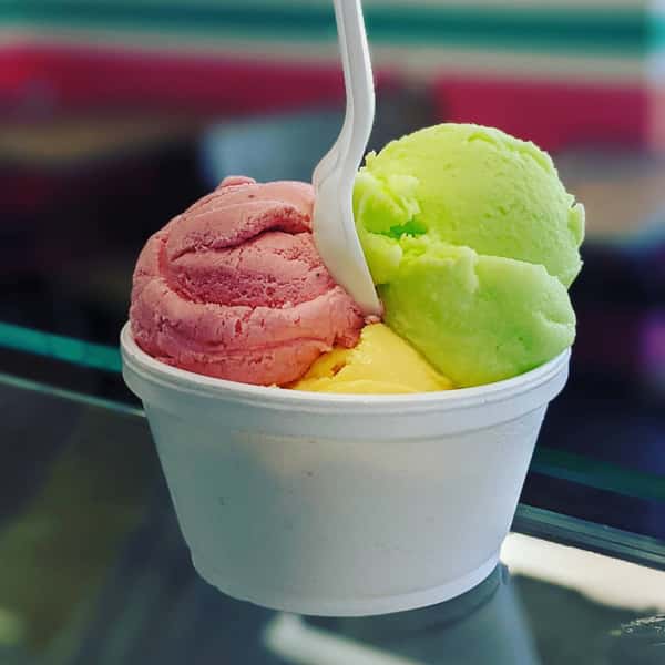 A cup of ice cream with three scoops: pink, yellow, and green, with a spoon in the center.