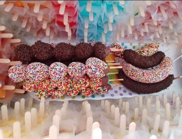 Colorful sprinkles cover chocolate-coated bananas and other treats on sticks, surrounded by bright candy and popsicle sticks.