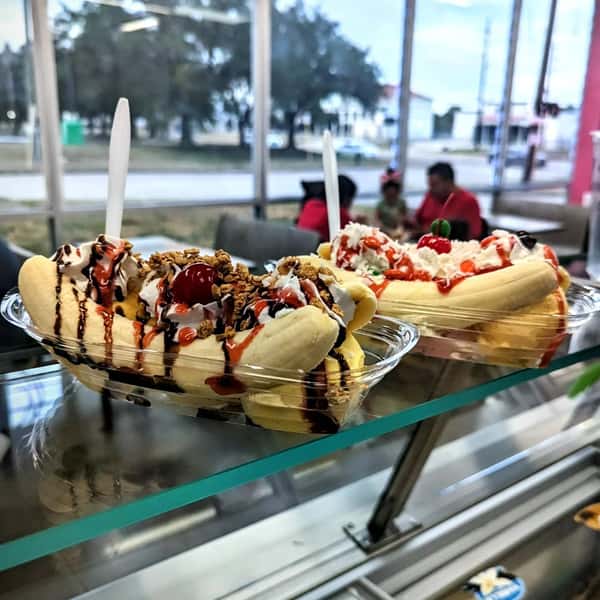 The image shows two banana splits topped with whipped cream, chocolate syrup, strawberry syrup, granola, and cherries.