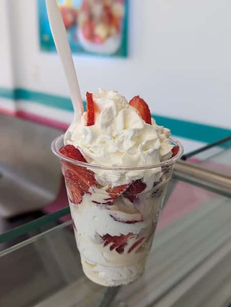A cup filled with layers of whipped cream, fresh strawberries, and creamy dessert, served with a spoon.