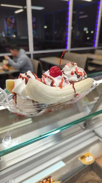 Banana split sundae topped with whipped cream, chocolate and strawberry syrup, and a cherry in a clear plastic dish.