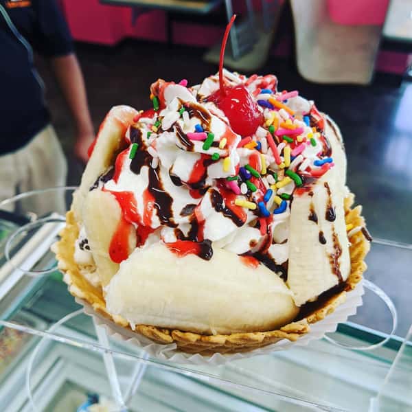 A banana sundae in a waffle bowl, topped with whipped cream, chocolate and strawberry syrup, sprinkles, and a cherry.