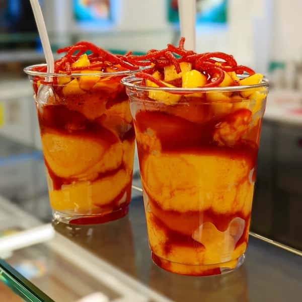 Two cups filled with mango, chamoy, and chili powder, topped with a spiral of tamarind candy and a drizzle of sauce.