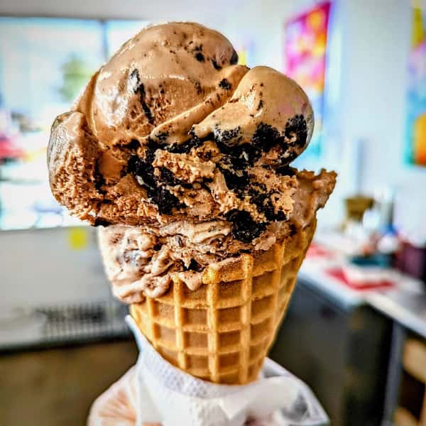 A cone topped with two scoops of chocolate ice cream mixed with cookie crumbs, with a blurred background of a shop setting.
