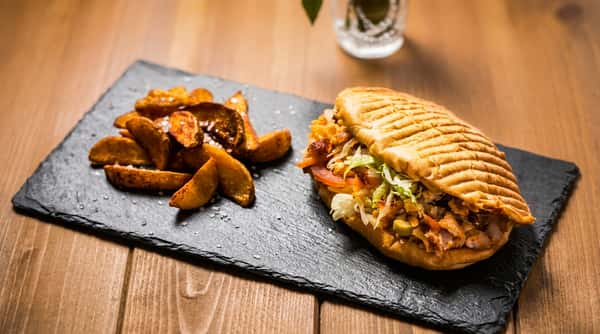 Sandwich with meat, lettuce, tomatoes, and red cabbage, served with potato wedges on a slate plate.