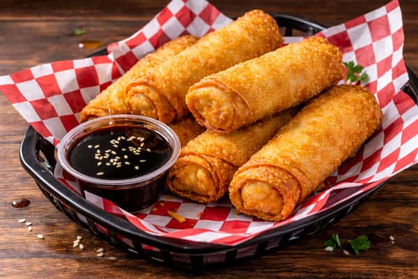 Golden crispy egg rolls served in a basket with soy dipping sauce and sesame seeds on red checkered deli paper.