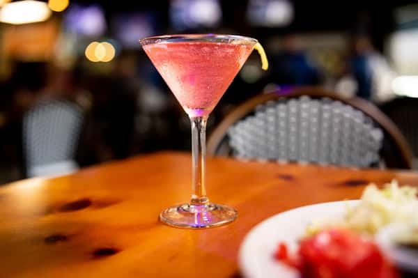 Chilled pink cocktail in a martini glass garnished with a lemon twist on a wooden table in a cozy, dimly lit restaurant.