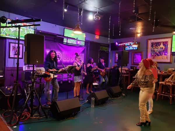Live band performing in a cozy bar with colorful lights, wall art, and patrons enjoying music and dancing near the stage.