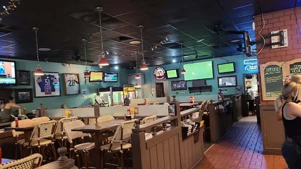 Casual sports bar dining area with multiple TVs, framed jerseys on teal walls, and cozy booth seating with wicker chairs.