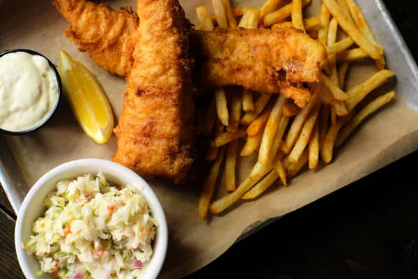 Crispy fried fish over golden fries with tartar sauce, lemon wedge, and a side of creamy coleslaw on a metal tray.