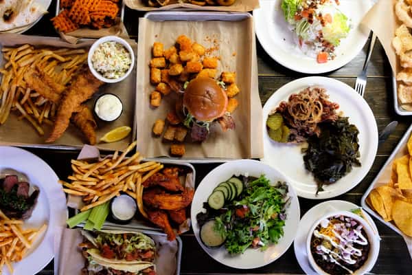 Assorted American comfort food including burgers, tacos, wings, fries, salad, and a variety of side dishes on a wooden table.