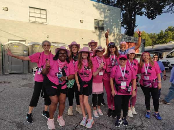 A fun team event in pink shirts and hats pose together, smiling and making peace signs, celebrating as a team outdoors.