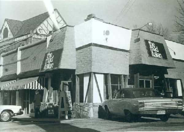 A vintage black-and-white photo of a restaurant building with signage reading "The Mad Italian." A classic car is parked nearby, with a striped awning visible on the left.