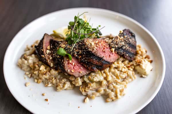Perfectly sliced steak, cooked to a tender medium-rare, is served over a hearty risotto or grain base. The dish is finished with a sprinkle of herbs and seeds, adding both crunch and visual appeal.