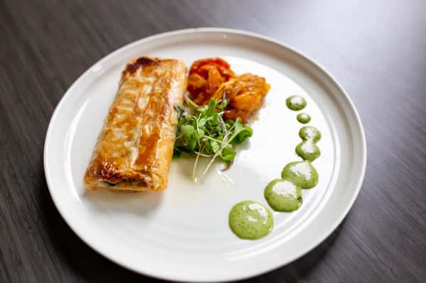 A beautifully baked salmon fillet with a light, flaky pastry or crisp topping sits at the center of the plate. It's paired with sautéed vegetables and a fresh herb garnish, complemented by a smooth green sauce artfully placed around the dish for a refined, restaurant-quality finish.