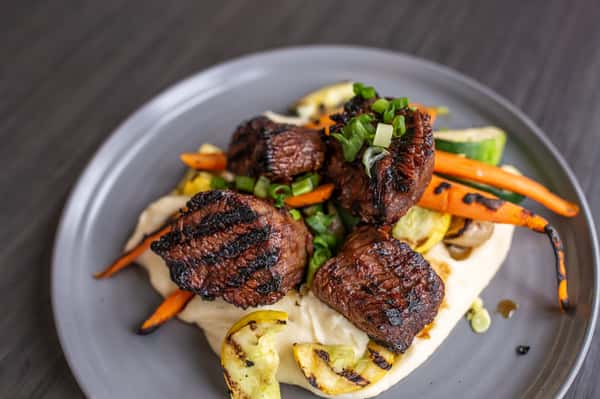 Grilled steak medallions with a deep, flavorful char are served alongside roasted vegetables and a grilled lemon, adding both color and a touch of citrus brightness.