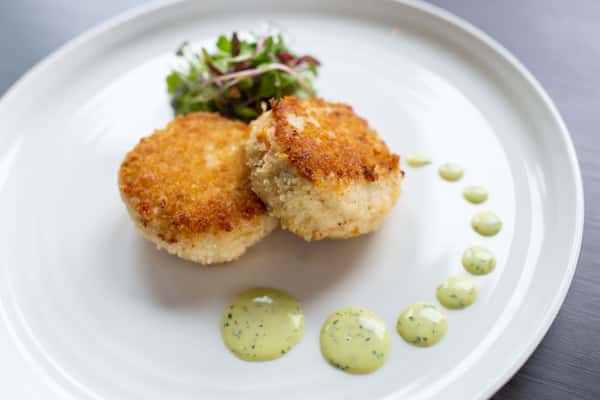 This dish features two golden, crispy-coated rounds—likely crab cakes or risotto cakes—perfectly seared for a crunchy exterior. They are served with a delicate green herb sauce dotted across the plate and finished with a small garnish of fresh greens, creating a clean and elegant presentation.