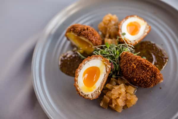 This image showcases a beautifully plated appetizer featuring two perfectly soft-boiled eggs with golden, jammy yolks, cut in half and coated in a crispy, golden crust. The eggs are paired with a rich dipping sauce and a side of what appears to be finely chopped or sautéed vegetables. Garnished with fresh herbs, the dish offers a striking contrast of textures—crispy on the outside, creamy on the inside—presented with a refined, modern touch.