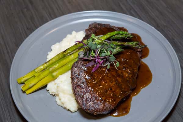 A juicy steak is served alongside tender asparagus and creamy mashed potatoes, all finished with a rich, savory sauce. The dish is hearty, classic, and presented with a polished, upscale touch.