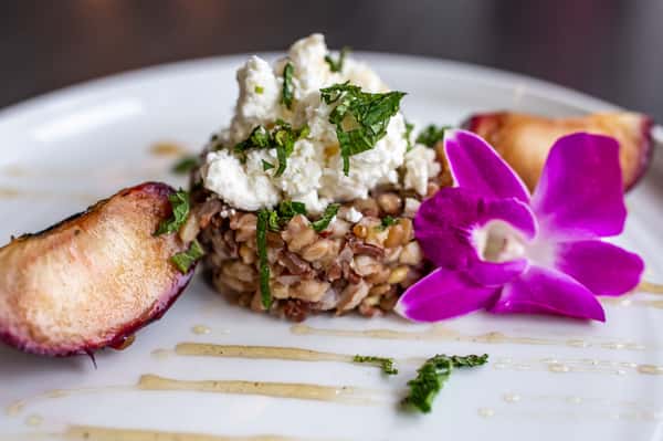 This image presents an elegant, chef-crafted dish featuring a hearty grain base—likely risotto or farro—blended with finely chopped vegetables. It's topped with crumbled white cheese that adds a creamy, slightly tangy contrast to the dish. A slice of toasted bread sits on the side, while a delicate drizzle of sauce enhances the presentation. The plate is finished with a vibrant purple orchid, adding a pop of color and a refined, upscale touch.