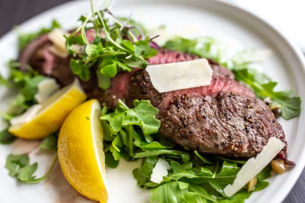 A perfectly cooked steak is sliced and served over a bed of fresh greens, topped with shaved cheese and microgreens. A bright lemon wedge adds a fresh, zesty touch to balance the rich flavors.