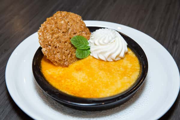 This image shows a rich crème brûlée with a golden, caramelized top served in a black dish. It's topped with whipped cream, a fresh mint leaf, and a crunchy nut-coated garnish. The combination of creamy, crisp, and fresh elements makes it look elegant and indulgent.