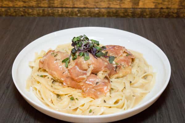 This image showcases a comforting and elegant pasta dish, featuring fettuccine noodles coated in a light, creamy sauce. Thin slices of salmon are layered on top, adding a delicate, savory richness to the plate. The dish is finished with a sprinkle of herbs and microgreens, bringing freshness and color to the presentation, making it both visually appealing and appetizing.