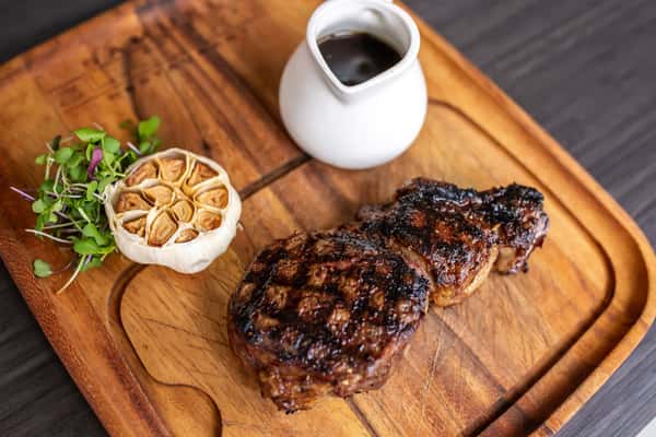 A grilled steak with a flavorful, charred exterior is plated alongside roasted vegetables and accompanied by a small serving of sauce. The dish is complemented by a small pitcher of jus, emphasizing its rich, savory profile and upscale presentation.
