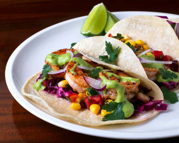 Shrimp tacos with red cabbage, corn, tomato, and green sauce on corn tortillas, garnished with cilantro and lime wedges.