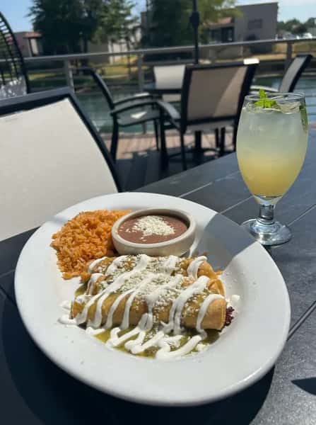 Mexican enchiladas topped with green sauce, crema, and queso, served with rice and beans alongside a refreshing citrus cocktail on a sunny patio