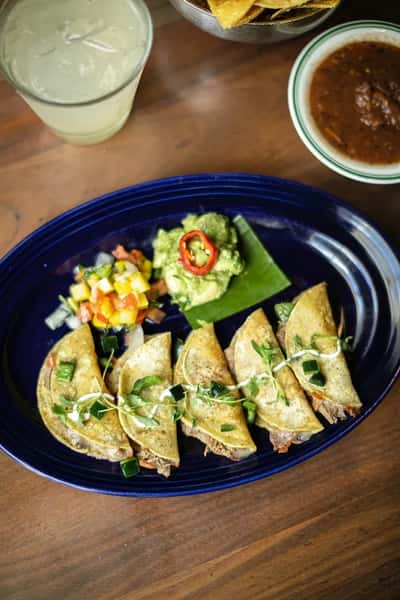Plated beef tacos topped with microgreens, served with guacamole, pico de gallo, tortilla chips, salsa, and a margarita on a rustic wooden table.