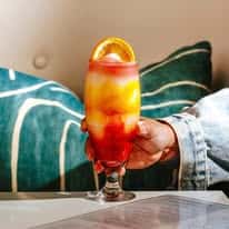 Alt text: Vibrant layered tropical cocktail with orange and red hues garnished with citrus, served in a tall glass at a modern restaurant lounge