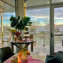 Bright waterfront restaurant interior with large windows, elegant table setting, and scenic outdoor patio views