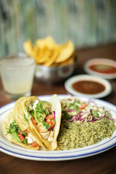 Mexican chicken tacos topped with avocado, pico de gallo, and cilantro, served with green rice and fresh cabbage slaw, alongside tortilla chips, salsa, and a margarita on a rustic restaurant table.