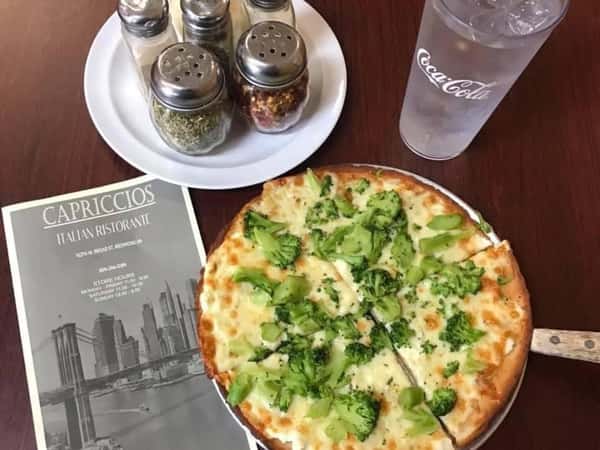 A golden-crust broccoli and cheese pizza on a dark wood table beside a Capriccios Italian Ristorante menu, spice shakers, and a glass of iced water.