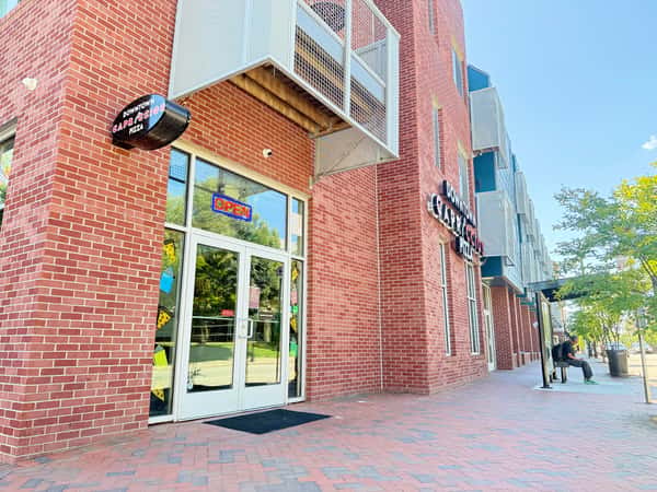 Exterior of Downtown Capriccios Pizza featuring a red brick facade, glass storefront with an "Open" sign, and a sidewalk leading to a bus stop.