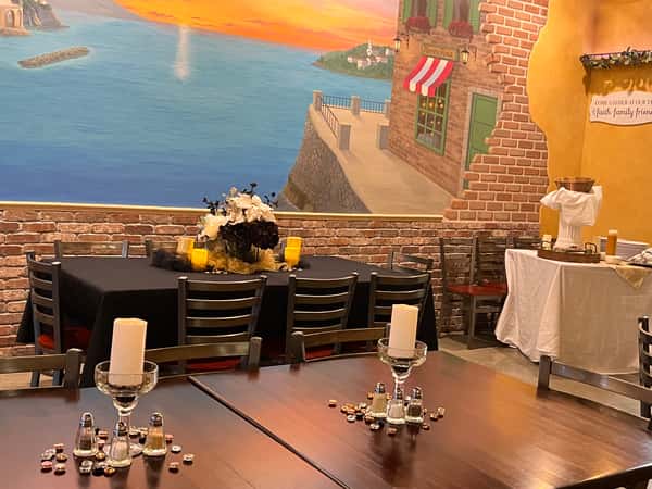 An inviting restaurant dining room features a large Mediterranean coastal mural, tables set with candles and floral centerpieces, and rustic brick accents.