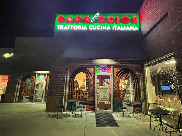 Vibrant red and green neon signs highlight the brick storefront of Capriccios Trattoria Cucina Italiana at night, featuring Italian landmark window graphics and outdoor seating.