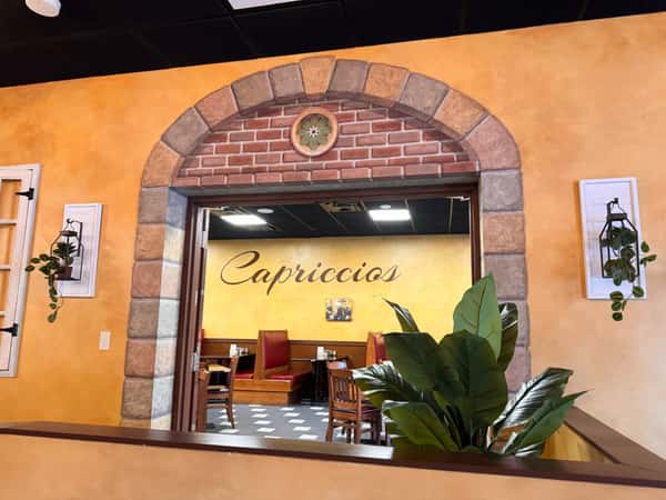 An inviting view through a rustic stone archway into Capriccios restaurant, featuring warm yellow walls, red booths, and lush green plants.
