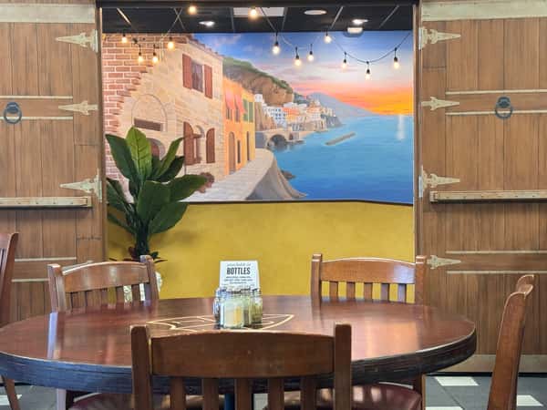 A round wooden dining table and chairs set in front of a colorful Mediterranean coastal sunset mural framed by large rustic wooden doors.