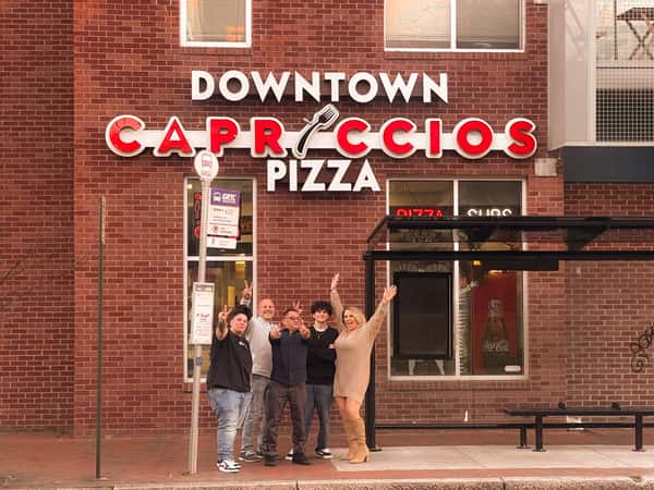 A group of diverse people posing happily in front of the Downtown Capriccios Pizza restaurant's red brick exterior and large illuminated sign.
