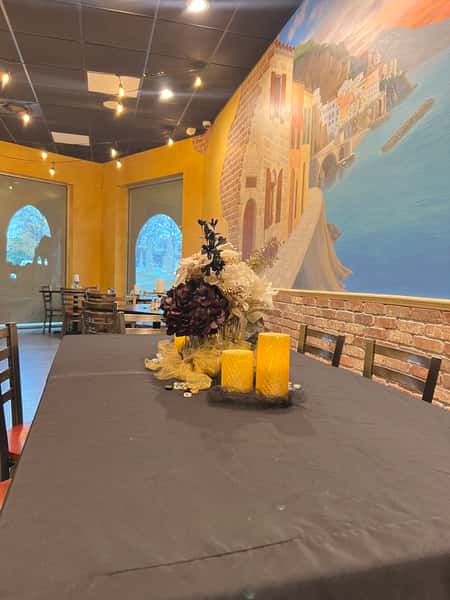 Restaurant dining room featuring a black-clothed table with a floral centerpiece and yellow candles against a colorful Mediterranean mural and yellow walls.