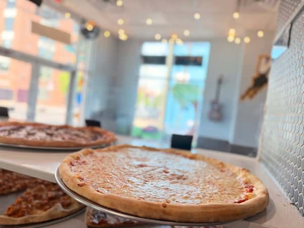 A close-up of a golden-brown cheese pizza on a metal tray in a bright, modern pizzeria with warm string lights in the background.