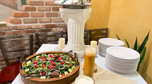 A fresh garden salad with red tomatoes and purple onions in a large wooden bowl on a buffet table with stacks of white plates and dressings.