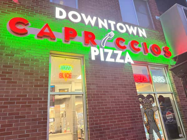 Nighttime storefront of Downtown Capriccios Pizza featuring vibrant red neon signage and a green glow against a brick wall.