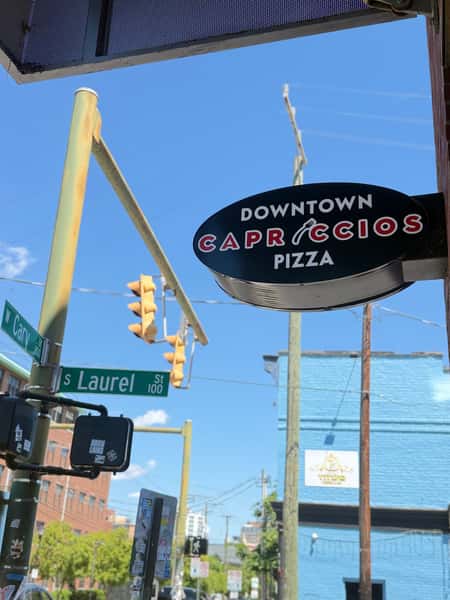 Downtown Capriccios Pizza oval black sign on an urban street corner with a clear blue sky and traffic lights.