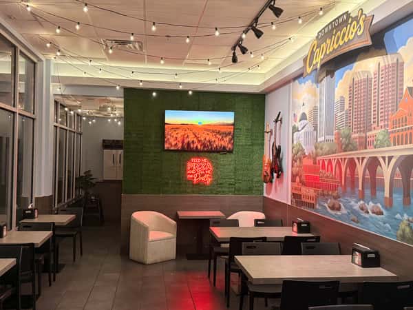 Modern restaurant interior featuring a colorful city mural, a green moss wall with a red neon sign, and warm string lights over dining tables.