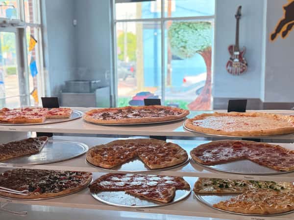 A variety of freshly baked pizzas on tiered white display shelves in a bright pizza shop with a guitar on the wall.